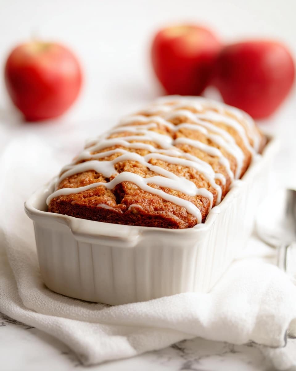 The image shows a golden brown baked cake with a rough textured top, drizzled with a smooth white glaze in an irregular criss-cross pattern. The cake is in a white rectangular ceramic dish with curved edges. The dish sits on a soft white cloth on a white marbled surface. In the blurred background, there are three red apples that add a fresh element to the scene. A silver spoon is partially visible in the foreground near the cloth. photo taken with an iphone --ar 4:5 --v 7