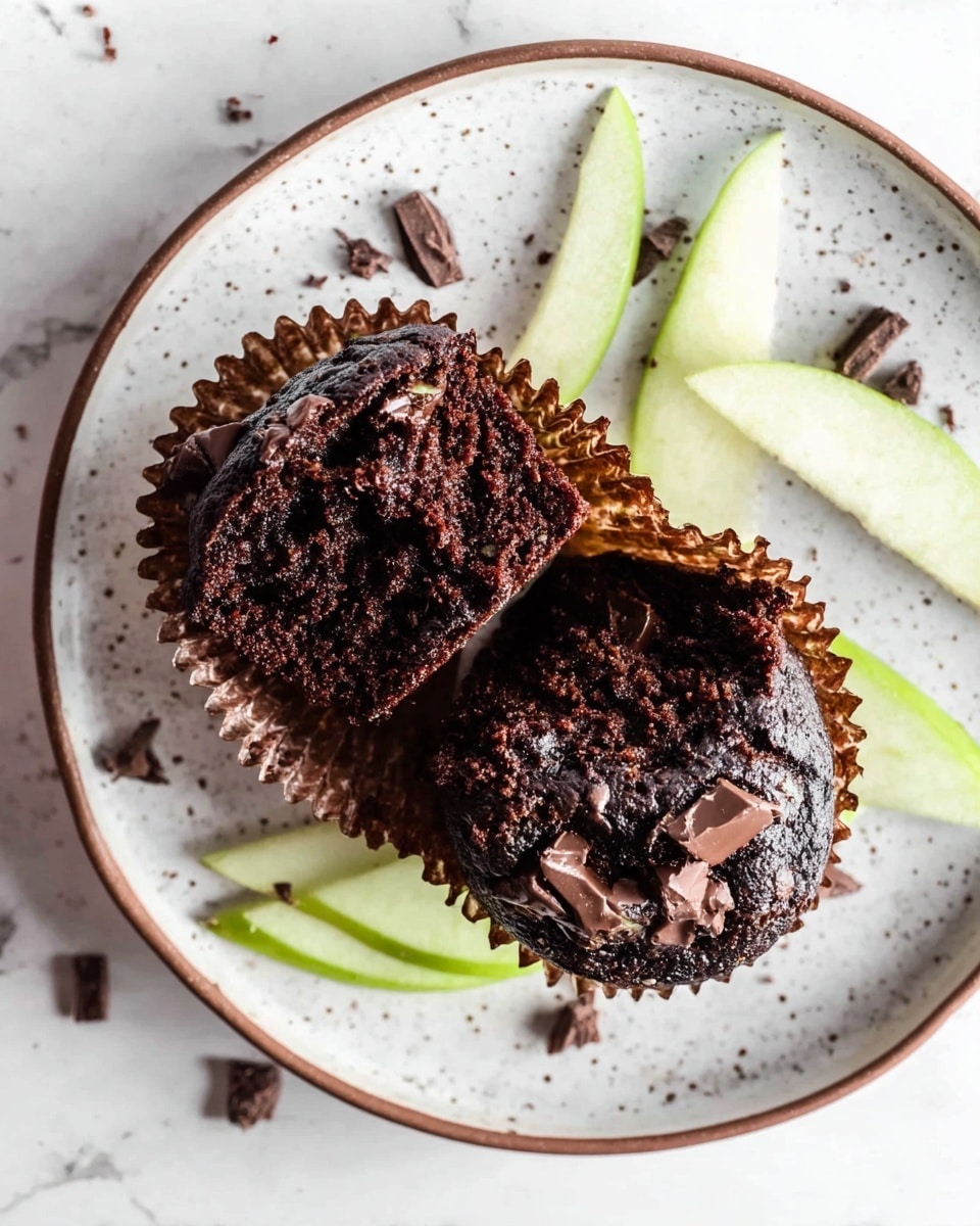 A white speckled plate with a brown rim holds two pieces of a dark chocolate muffin, broken in half to show their moist, rich black-brown interior with melted chocolate chunks scattered throughout. The muffin wrappers, dark brown in color, are peeled back and spread out beneath the muffins on the plate. Thin light green apple slices are scattered around the muffins and plate. The scene is set against a white marbled texture background. Photo taken with an iphone --ar 4:5 --v 7