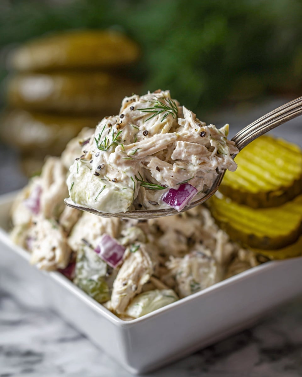 A close-up view of a creamy chicken salad held by a vintage silver spoon above a white square bowl filled with the same salad. The salad has shredded white chicken mixed with small chunks of purple-red onion, green dill, and flecks of black pepper in a thick white creamy dressing. At the back of the bowl, a small stack of golden yellow, slightly curved pickle slices lean against the salad. The background is softly blurred, showing a hint of green and a rustic setting. The photo is taken on a white marbled surface. Photo taken with an iphone --ar 4:5 --v 7