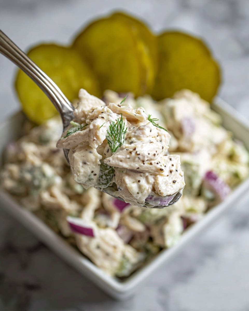 A close-up view of a spoonful of creamy chicken salad held above a white square bowl filled with more of the salad. The chicken salad has three visible layers: shredded white chicken mixed with small diced pieces of purple onion and green dill, all coated with a thick, white creamy dressing speckled with black pepper. In the background, several yellow-green pickle slices rest on top of the salad inside the bowl. The scene is set on a soft white marbled surface, giving a clean and fresh look. Photo taken with an iphone --ar 4:5 --v 7