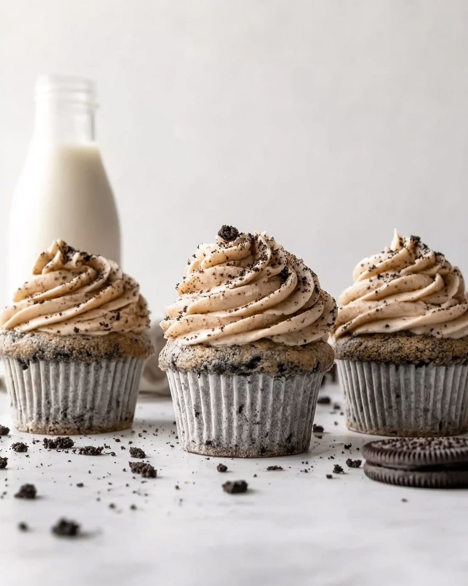 Three cookies and cream cupcakes are placed side by side on a white marbled surface, each with a light gray cupcake base speckled with dark cookie crumbs. Each cupcake is topped with a swirl of creamy, light brown frosting that has flecks of cookie bits, with the frosting rising in a soft peak. Small cookie crumbs are scattered over the surface in front of the cupcakes, and a glass bottle filled with milk is visible in the background against a plain white wall. photo taken with an iphone --ar 4:5 --v 7