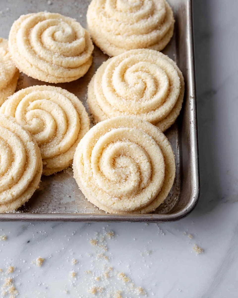 A group of eight swirl-shaped cookies are arranged closely in two rows on a silver baking tray, each cookie having a pale golden color with a soft crumbly texture visible on their ridges. They have a spiral pattern with distinct curved lines that start from the center and widen outward, giving each cookie a layered look. Small crumbs are scattered around the cookies on the tray, adding a sense of freshness. The background is a white marbled texture that contrasts gently with the tray and the cookies. Photo taken with an iphone --ar 4:5 --v 7