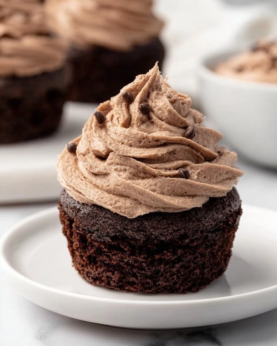 A single chocolate cupcake sits on a white plate, showing two main layers. The bottom layer is a dark, rich chocolate cake with a moist and slightly crumbly texture. On top is a thick swirl of light brown chocolate frosting, piped in a spiral with sharp ridges and a few small chocolate flecks visible throughout. The background features a white marbled surface, with blurred hints of more cupcakes and a bowl of the same frosting in the distance. Photo taken with an iphone --ar 4:5 --v 7