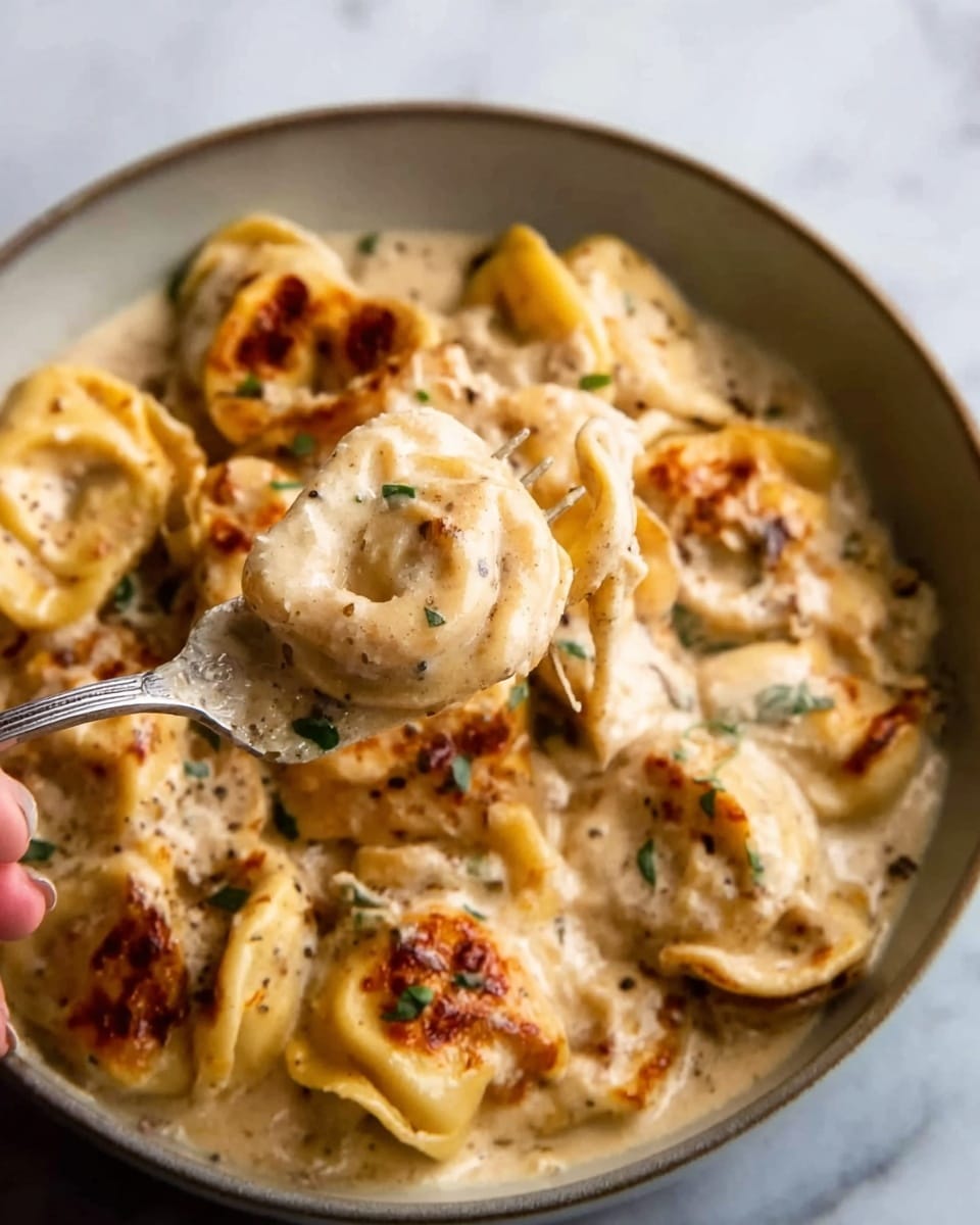 A close-up of a creamy pasta dish with several layers of tortellini covered in a thick, light beige sauce with visible bits of herbs and small green flecks, a woman's hand holding a silver fork lifts a portion from the center, showing browned golden-baked spots on the pasta, in a white bowl on a white marbled surface. photo taken with an iphone --ar 4:5 --v 7