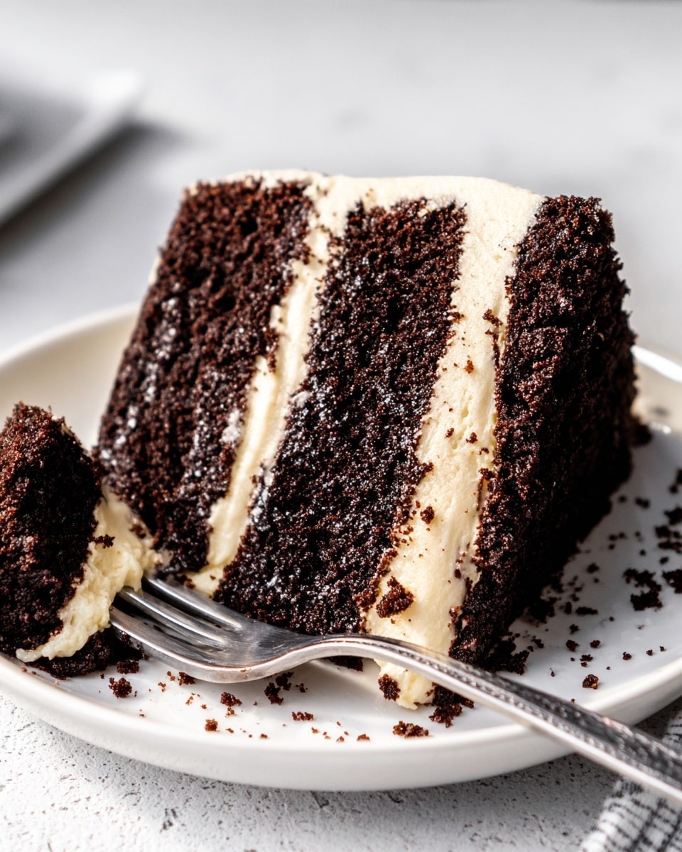 A close-up of a two-layer dark chocolate cake slice with moist, crumbly texture, separated by a thick, smooth, light beige cream layer. The cake layers have a rough surface showing crumbs, and the cream layers are even and soft-looking. A small piece of the cake has been cut and placed on a silver fork, showing the same two layers. The cake slice sits on a white plate with a few crumbs around, all set on a white marbled surface. Photo taken with an iphone --ar 4:5 --v 7