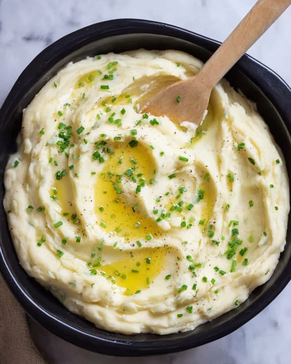 The image shows a black bowl filled with creamy, smooth mashed potatoes. The mashed potatoes have a white, soft texture with a few small lumps visible, giving it a natural, homemade look. There are small green chive pieces sprinkled across the top, adding a touch of color. A drizzle of golden melted butter sits on top, creating shiny, glossy spots. A wooden spoon is partially dipped into the mashed potatoes on the right side. The bowl is placed on a white marbled surface. Photo taken with an iphone --ar 4:5 --v 7