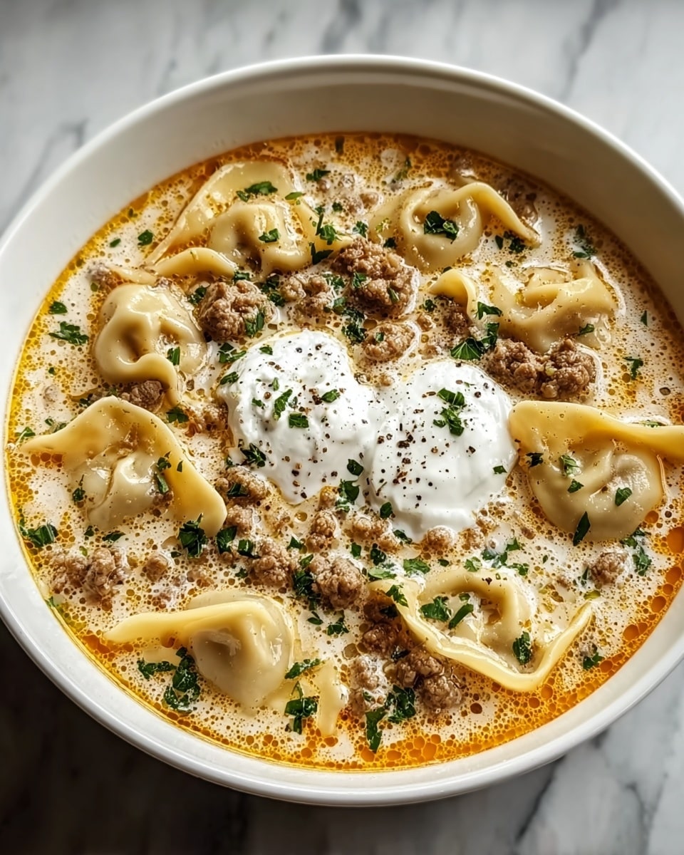 A white bowl filled with creamy soup that has a light yellow color with small orange oil spots on top. Floating in the soup are several folded pasta pieces that have a smooth and slightly wavy texture. Scattered crumbled browned ground meat pieces cover the surface along with green chopped herbs. In the center, two dollops of white sour cream sit, sprinkled with black pepper. The bowl is placed on a white marbled surface. photo taken with an iphone --ar 4:5 --v 7
