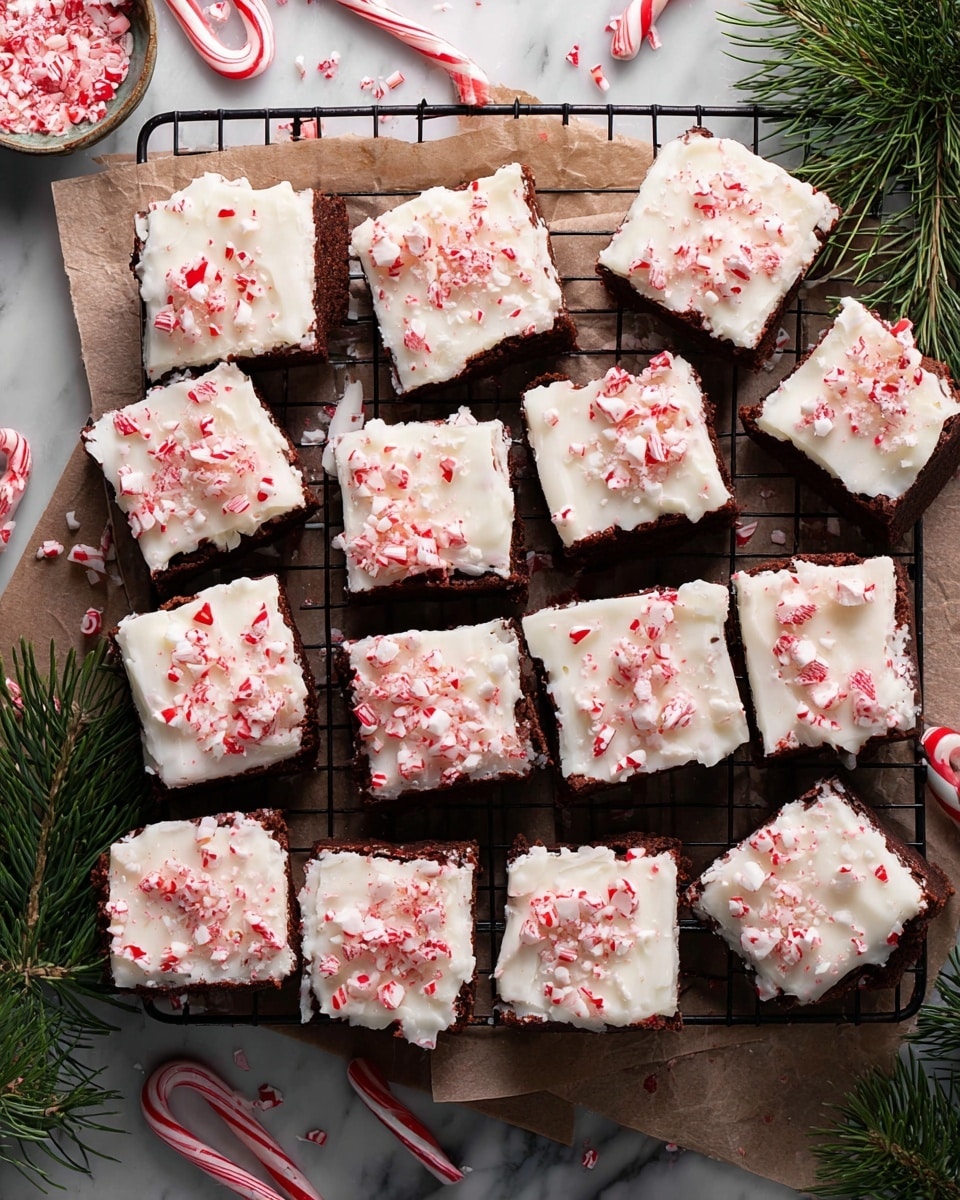 A grid of sixteen square pieces of brownies is shown on a black cooling rack lined with brown parchment paper. Each brownie has two layers: a dark brown, dense-looking chocolate base topped with a thick, white frosting layer with a creamy and smooth texture. The frosting is generously covered with small, uneven red and white crushed peppermint candy pieces, scattered all over the top surface. Around the rack are broken peppermint sticks, loose crushed candy, and some green fir sprigs, adding a festive touch. The whole scene is set on a white marbled textured surface. Photo taken with an iphone --ar 4:5 --v 7