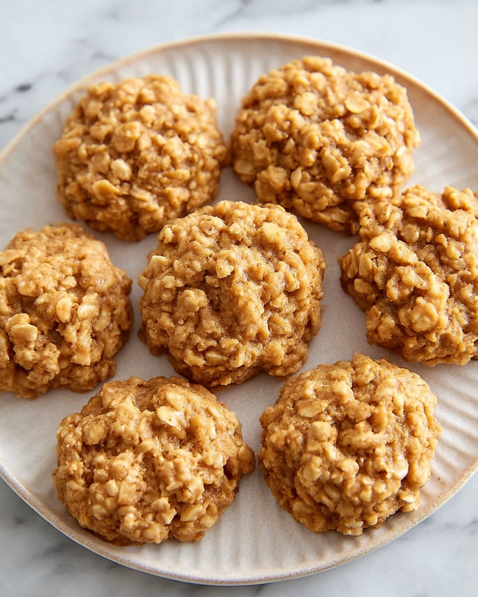 The image shows nine small oatmeal cookies placed evenly on a white plate with a textured edge. Each cookie is thick and round, with a rough surface filled with visible oats, giving them a chunky texture. The cookies are a light golden brown color, suggesting they are soft and chewy. The plate sits on a white marbled surface that adds a clean and bright background to the image. photo taken with an iphone --ar 4:5 --v 7