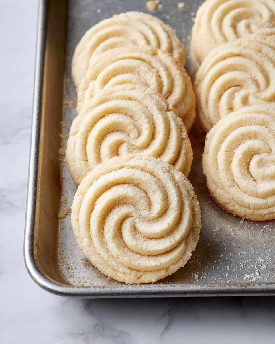 The image shows a group of light beige round cookies arranged in two neat rows on a metal tray, each cookie having a raised, spiral swirl pattern on top, making them look soft but firm with crumbly edges. The metal tray rests on a surface with a white marbled texture, and some cookie crumbs are scattered around them, giving a fresh-baked feel. Photo taken with an iphone --ar 4:5 --v 7
