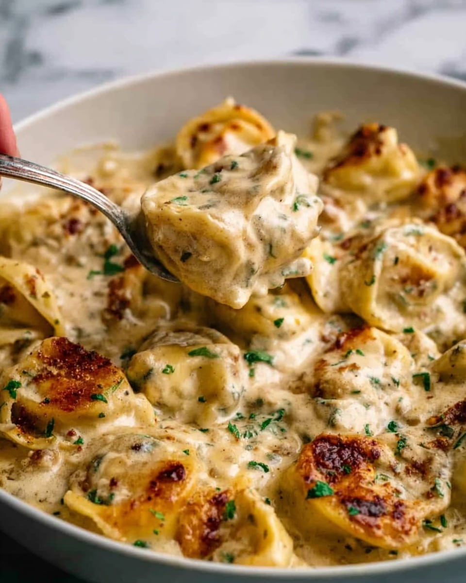 The image shows a close-up of a creamy pasta dish with several layers visible. The base layer consists of small tortellini pasta pieces in a pale yellow color, covered generously with a thick, creamy white sauce that has a smooth texture. Scattered throughout are small bits of crispy, browned bacon adding a reddish-brown, slightly rough texture on top. A woman's hand holding a white fork is lifting some of the tortellini, showing the pasta’s soft, pillowy surface and the rich sauce clinging to it. The dish sits on a clean white plate placed on a white marbled surface. The lighting highlights the glossy, creamy sauce and the golden-brown bits of bacon, making the dish look warm and inviting. Photo taken with an iphone --ar 4:5 --v 7