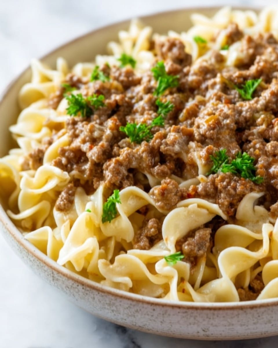 A close-up view of a white bowl filled with cooked egg noodles mixed with small pieces of browned ground meat, topped with small green parsley leaves scattered evenly across the surface. The noodles are thick, curly, and creamy in color, while the meat pieces are browned and crumbly, creating a hearty texture contrast. The image shows a soft focus on the noodles with a slight shine, highlighting their smooth texture. The bowl is placed on a white marbled surface. photo taken with an iphone --ar 4:5 --v 7