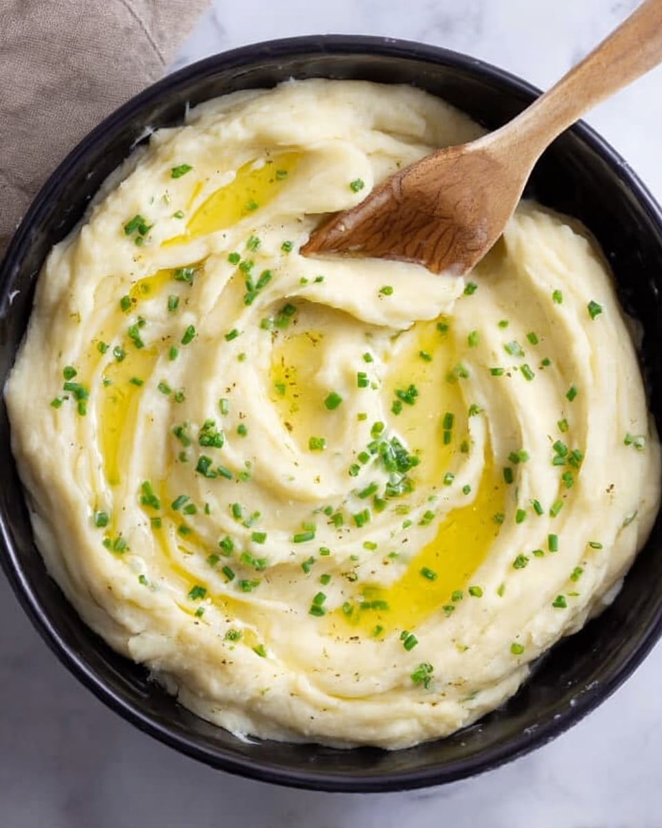 The image shows a black bowl filled with creamy mashed potatoes that have a smooth, fluffy texture. The top layer is swirled gently with a wooden spatula resting inside the bowl. There is a small pool of melted butter creating a golden-yellow shine on the surface, and green chopped chives are sprinkled evenly over the potatoes, adding a pop of color. The bowl is placed on a white marbled surface. Photo taken with an iphone --ar 4:5 --v 7