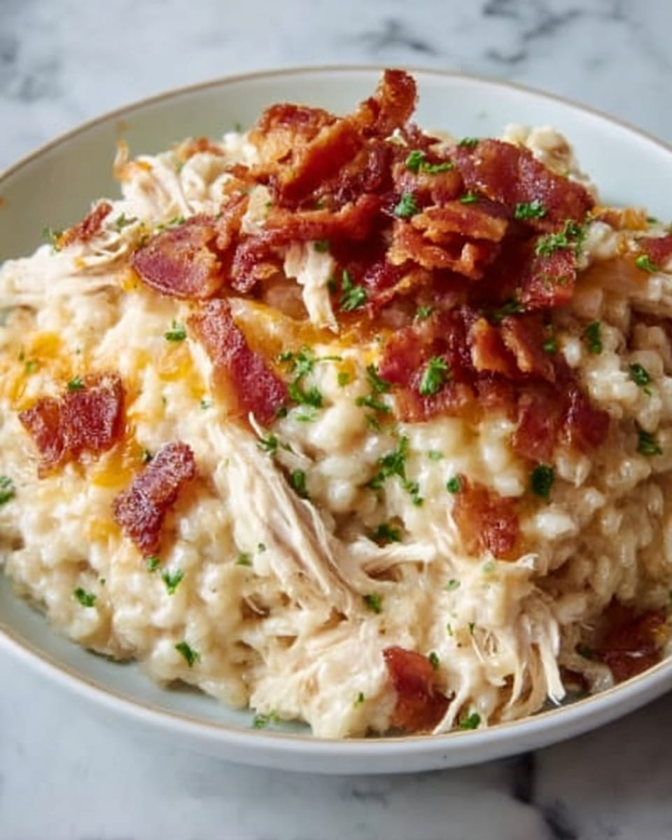 The image shows a close-up of a creamy chicken and rice dish on a white plate. The bottom layer is a soft, light beige rice mixed with small pieces of shredded chicken. On top, there is melted cheese, golden and bubbly, covering the chicken and rice. There are small, crispy, reddish-brown bacon bits sprinkled over the cheese, adding texture and color. Tiny green parsley pieces are scattered on the surface for a fresh look. The whole dish looks warm and gooey, with the melted cheese stretching slightly. The background is a white marbled texture. Photo taken with an iphone --ar 4:5 --v 7