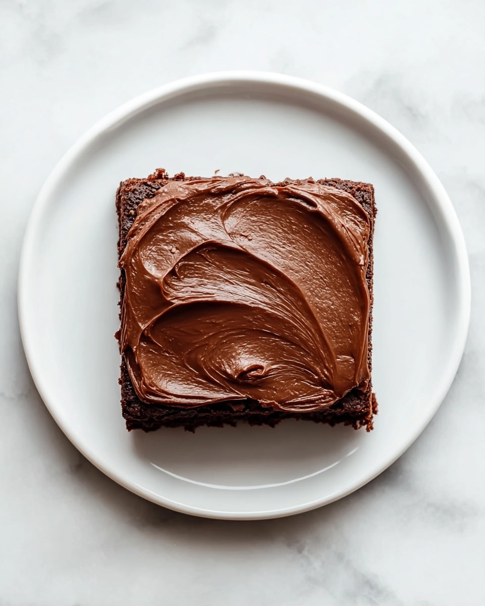 A single square piece of chocolate brownie sits in the center of a white round plate, placed on a white marbled surface. The brownie has a thick, rich layer of smooth, glossy dark chocolate frosting spread evenly across the top, with visible swirls and soft peaks giving it a textured look. The edges of the brownie are slightly crisp and crumbly, contrasting with the creamy frosting. The top-down view emphasizes the neat, clean shape and the inviting shine of the chocolate frosting. photo taken with an iphone --ar 4:5 --v 7