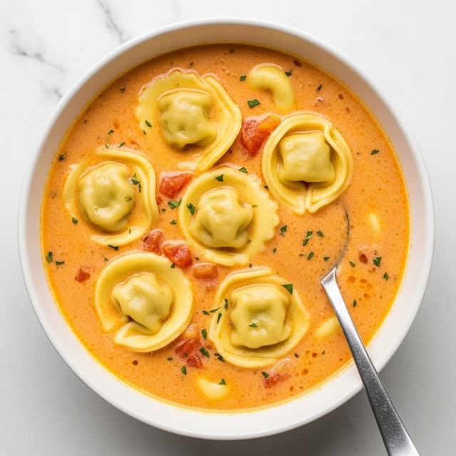 A white bowl filled with creamy yellow soup that has a smooth texture and small bits of orange vegetables inside. Floating on top are six round tortellini with a soft, folded shape, slightly lighter yellow than the soup. The soup is sprinkled with small green herb pieces and black pepper. A silver spoon is placed inside the bowl near the top right edge. The background is a white marbled texture. photo taken with an iphone --ar 4:5 --v 7