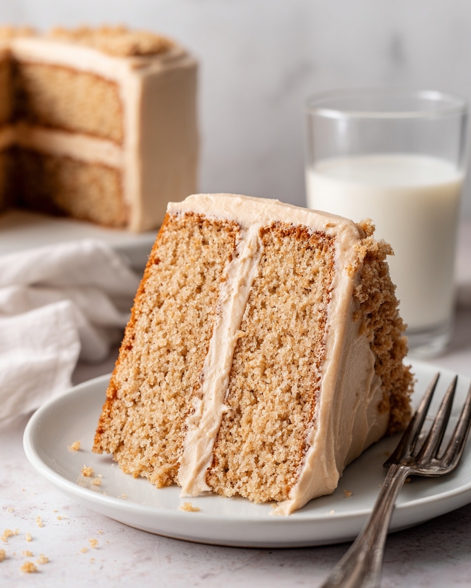 A slice of two-layer light brown cake sits on a white plate, with a thick layer of creamy, smooth, light brown frosting covering the top and sides. Between the two cake layers is an additional layer of frosting with small crumble pieces embedded. The cake texture looks moist and fluffy, and some frosting with crumbles decorates the side. Behind the plate is a clear glass filled with white milk on a white marbled texture, and a silver spoon lies in front of the plate. photo taken with an iphone --ar 4:5 --v 7