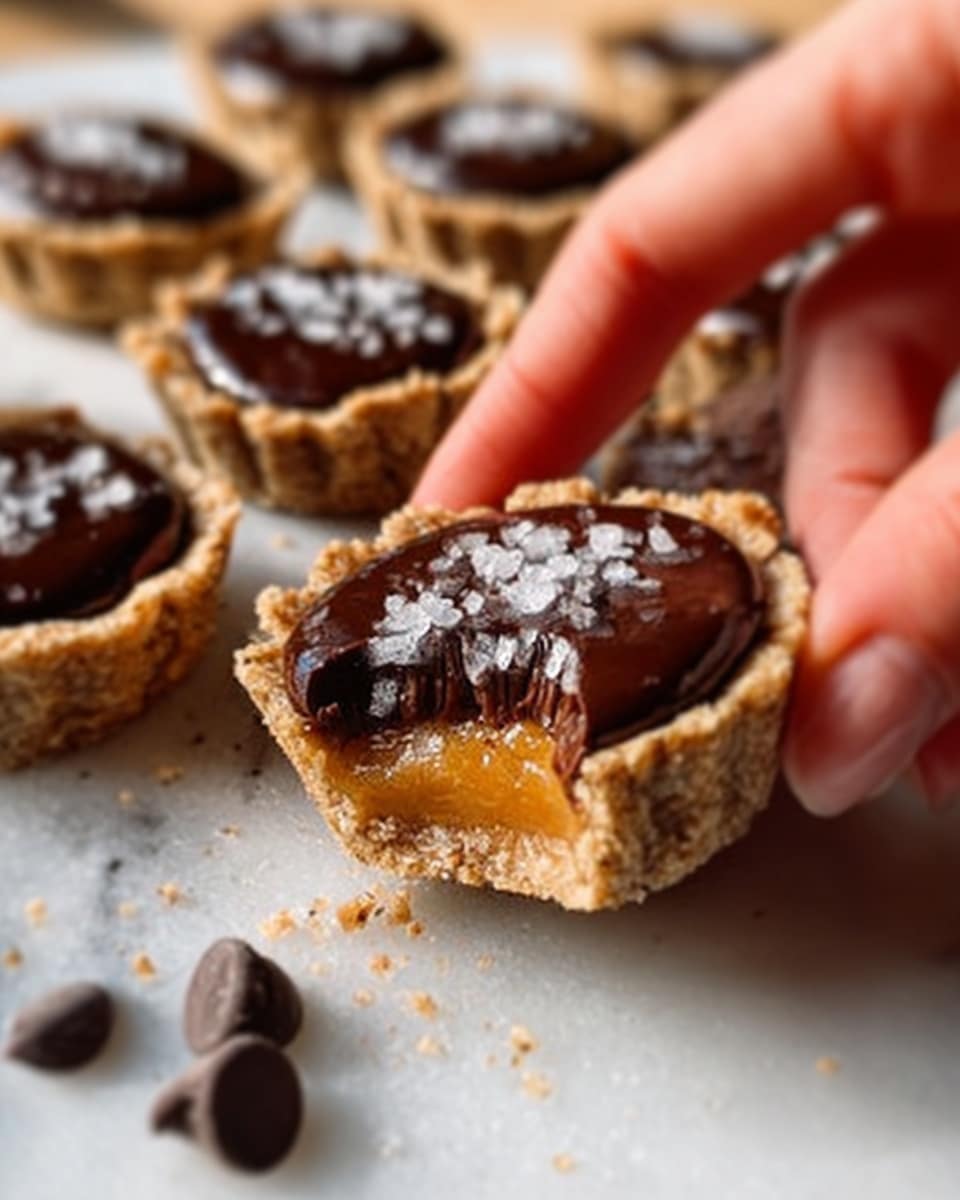 The image shows a woman's hand holding a small round dessert made of three visible layers. The bottom layer is a rough, crumbly crust in light brown color, the middle layer is a smooth golden caramel, and the top layer is a shiny, dark brown chocolate glaze. A few salt flakes are sprinkled on top of the chocolate layer. Several similar desserts are placed on a white marbled surface around the main focus, with soft sunlight casting gentle shadows. Photo taken with an iphone --ar 4:5 --v 7