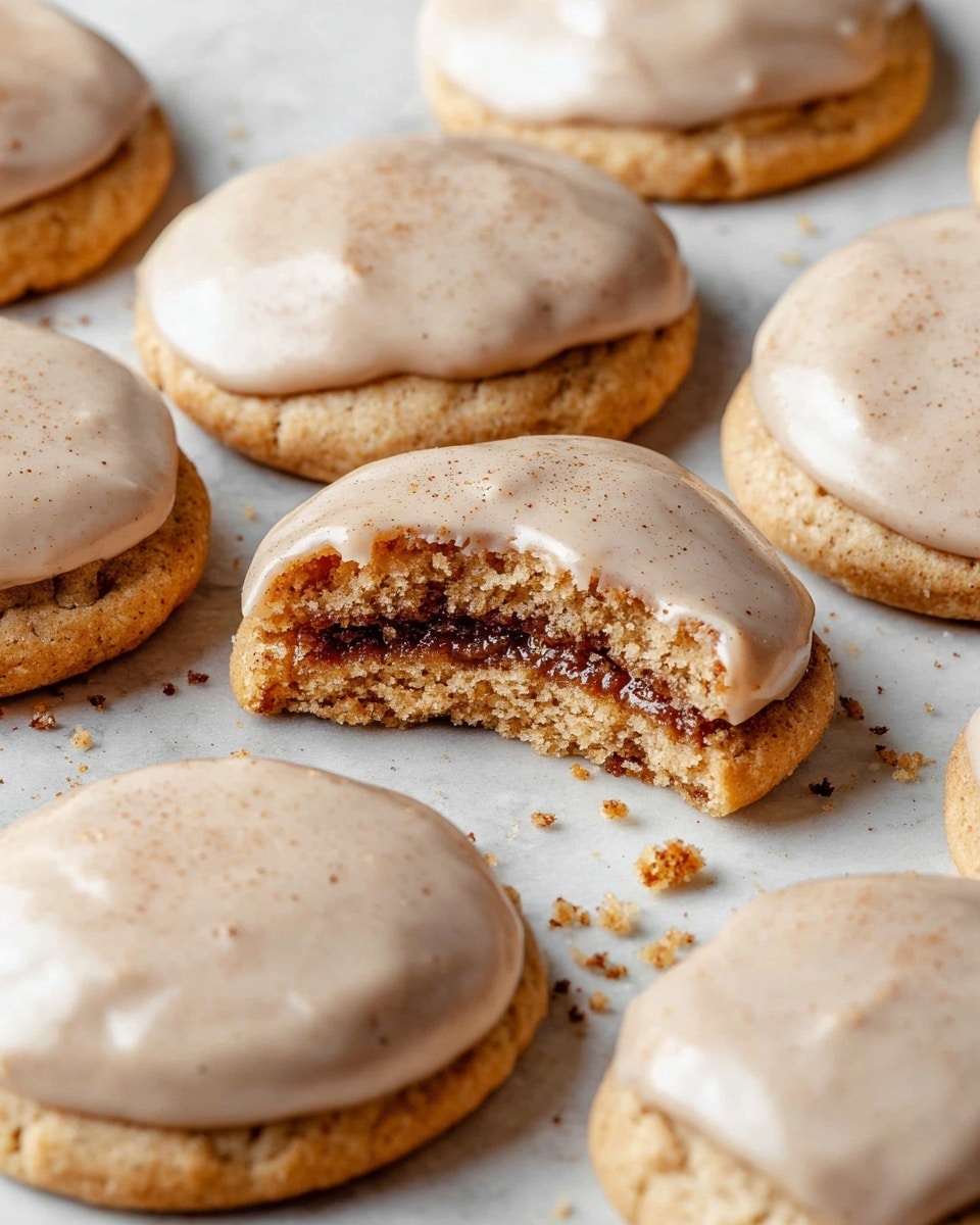 A group of round cookies with a light golden-brown base are shown on a white marbled surface, each topped with a smooth, light beige glaze that has a slight sheen and specks of darker color throughout. One cookie in the center is broken in half, revealing a soft, fluffy interior with a dark cinnamon filling inside the middle layer. Small crumbs are scattered around the cookies, adding texture to the scene. The cookies have a soft, slightly thick edge with the glaze evenly spread on top. Photo taken with an iphone --ar 4:5 --v 7