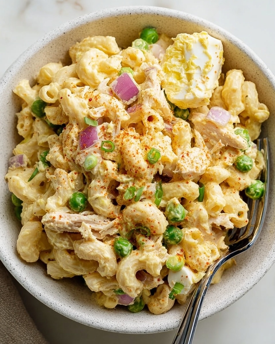 A close-up view of a bowl filled with creamy macaroni salad. The base layer shows yellowish macaroni elbows coated in a thick, pale yellow mayonnaise dressing. Scattered throughout are chunks of pale cooked chicken, small pieces of boiled egg with white and yellow layers, green peas, small diced purple onion bits, and green chopped scallions. There is a light sprinkle of red paprika or chili powder on top, adding a hint of color contrast. The bowl is white with small speckles, and a silver fork with a black handle rests on the right edge of the bowl. The background has a white marbled texture. photo taken with an iphone --ar 4:5 --v 7