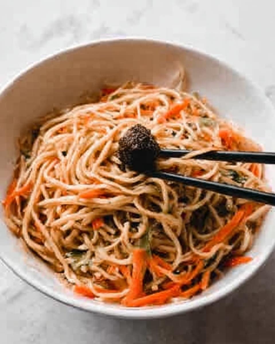 The image shows a close-up of a bowl filled with noodles mixed with thin orange carrot strips and small green herbs. On top, a pair of black chopsticks holds some noodles, lifting them from the bowl. The noodles are light beige, slightly shiny from sauce, with a soft texture. The bowl is white and sits on a white marbled surface, giving a clean, simple look. Photo taken with an iphone --ar 4:5 --v 7
