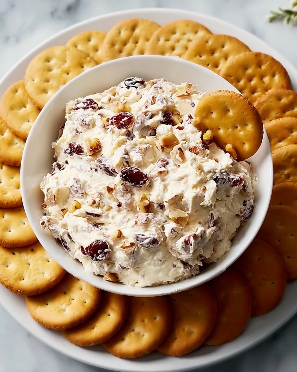 A white bowl filled with a creamy, thick spread mixed with chopped nuts and dried cranberries, giving it a textured look with white, brown, and dark red colors unevenly distributed. A round golden-brown cracker is slightly inserted into the spread on the right side of the bowl. The bowl sits in the center of a white plate covered with a neat circle of similar golden-brown crackers around it. The surface underneath is white marble, visible around the plate edges. photo taken with an iphone --ar 4:5 --v 7