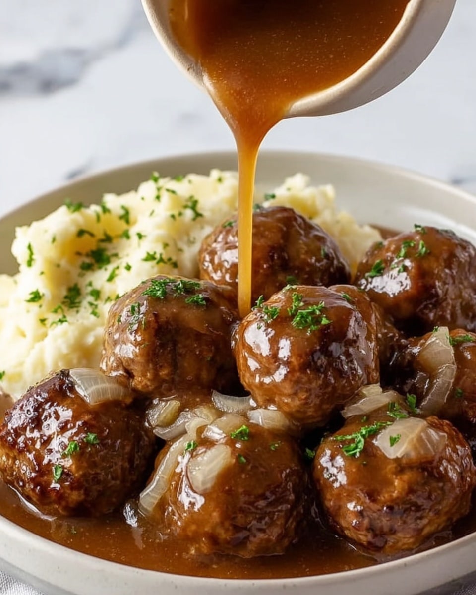 The image shows a white plate filled with eight brown meatballs covered in a glossy, rich brown gravy with visible cooked onion slices. The gravy is being poured over the meatballs, creating a shiny, wet texture. The meatballs are garnished with small bits of green herbs sprinkled on top. On the side of the plate, there is a creamy, crumbly mashed potato portion also sprinkled with green herbs. The plate is set against a white marbled surface. photo taken with an iphone --ar 4:5 --v 7