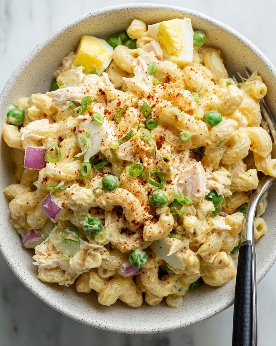 A close-up view of a creamy macaroni salad served in a white bowl with a speckled texture, placed on a white marbled surface. The salad has three visible layers: the first is a base of elbow macaroni pasta coated in a light yellow mayonnaise dressing, the second layer is mixed with chunks of boiled egg and pieces of tender chicken, and the third layer includes bright green peas and finely chopped red onions scattered throughout. The dish is topped with a light dusting of paprika and small sprinkles of chopped green onions. A silver fork with a black handle rests inside the bowl on the right side. Photo taken with an iphone --ar 4:5 --v 7