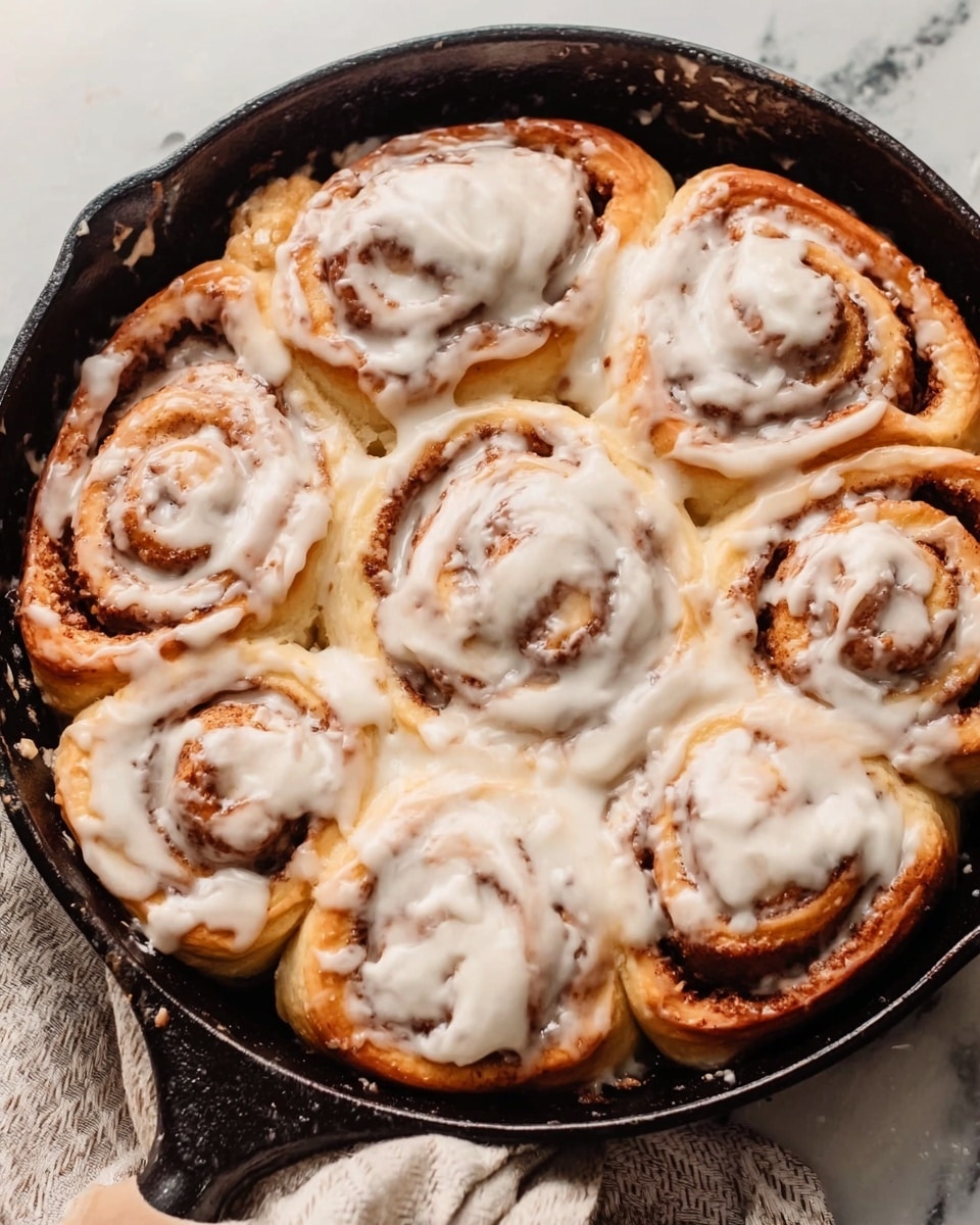 The image shows a cast iron pan filled with eight cinnamon rolls covered in a smooth, creamy white icing. Each roll is thick, golden brown with visible spiraled layers of cinnamon inside. The icing is spread evenly, slightly melting over the edges of the soft rolls, creating a shiny, moist look. The pan is placed on a white marbled surface, with a striped cloth partially visible on the side. photo taken with an iphone --ar 4:5 --v 7