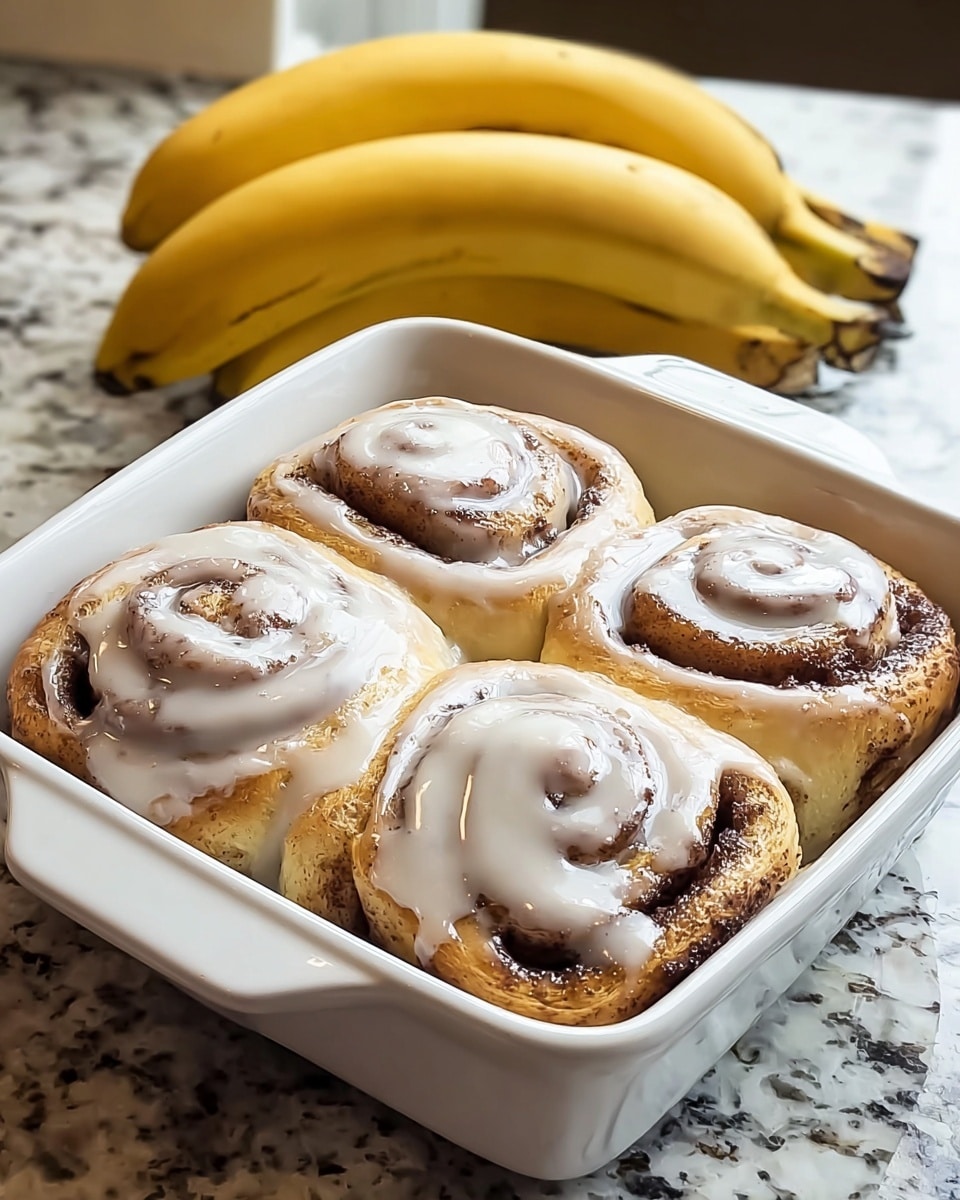 The image shows four cinnamon rolls tightly placed in a white square baking dish. Each roll has a soft golden-brown color with a spiral pattern of darker cinnamon filling visible through the dough. A thick, creamy white icing is generously drizzled over the top, glistening and slightly dripping down the sides of the rolls. The background is a white marbled textured countertop with a bunch of yellow bananas resting behind the dish. Photo taken with an iphone --ar 4:5 --v 7