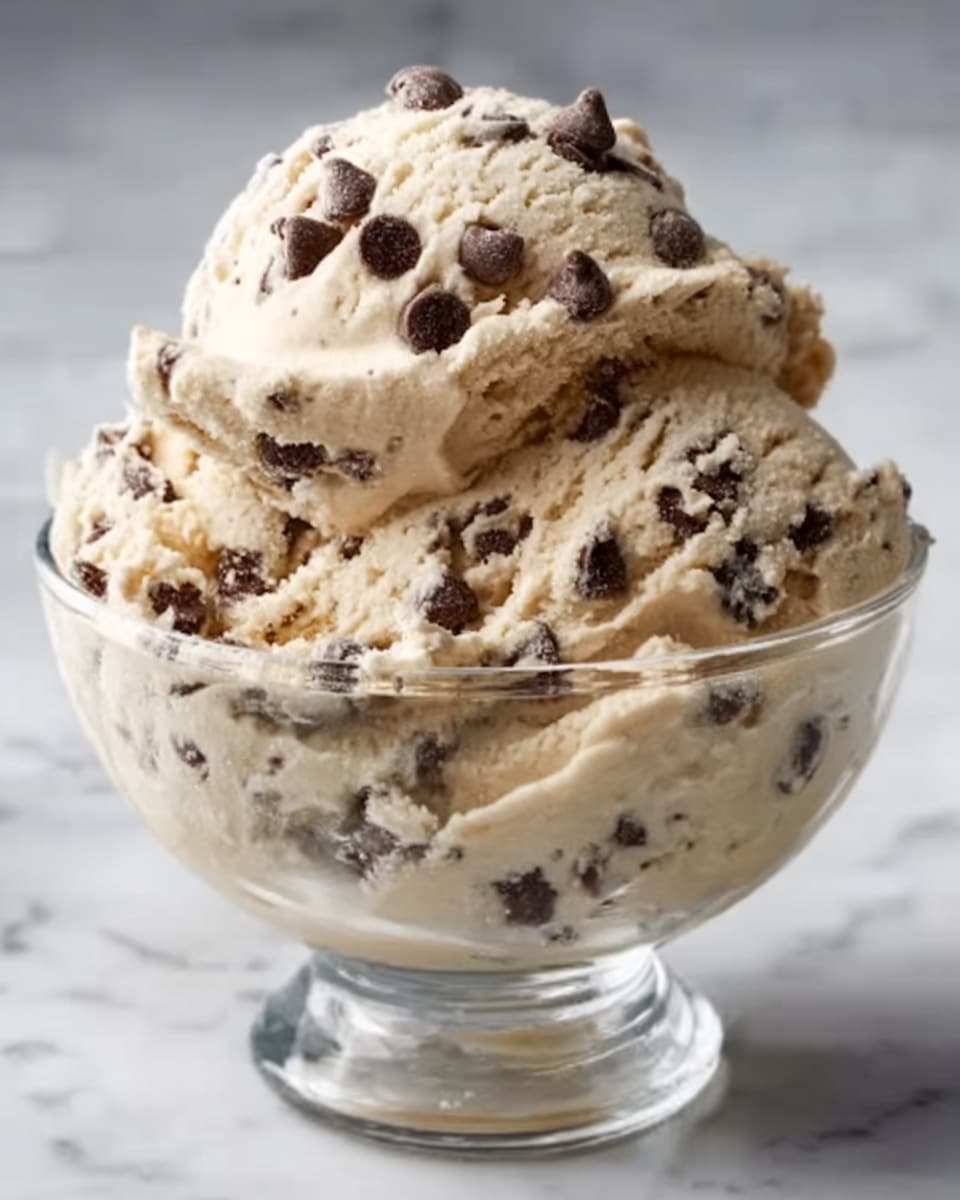 A clear glass bowl filled with two thick layers of creamy light brown ice cream mixed with dark chocolate chips spread evenly throughout. The ice cream is slightly scooped at the top, forming soft peaks with more chocolate chips scattered generously on the surface. The bowl sits on a white marbled background, highlighting the smooth texture and rich color contrast of the ice cream. photo taken with an iphone --ar 4:5 --v 7