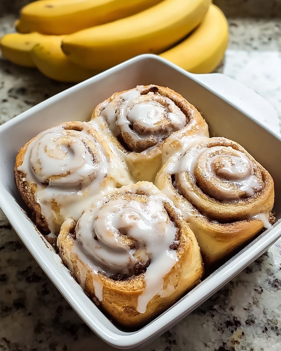 A white square ceramic dish holds four cinnamon rolls arranged in a two-by-two grid. Each roll has a visible swirl shape covered with a layer of thick, glossy white icing that drips slightly down the sides. The cinnamon rolls are golden brown with darker cinnamon filling visible in the spiral design. The dish sits on a white marbled surface with a bunch of ripe yellow bananas in the blurred background. Photo taken with an iphone --ar 4:5 --v 7