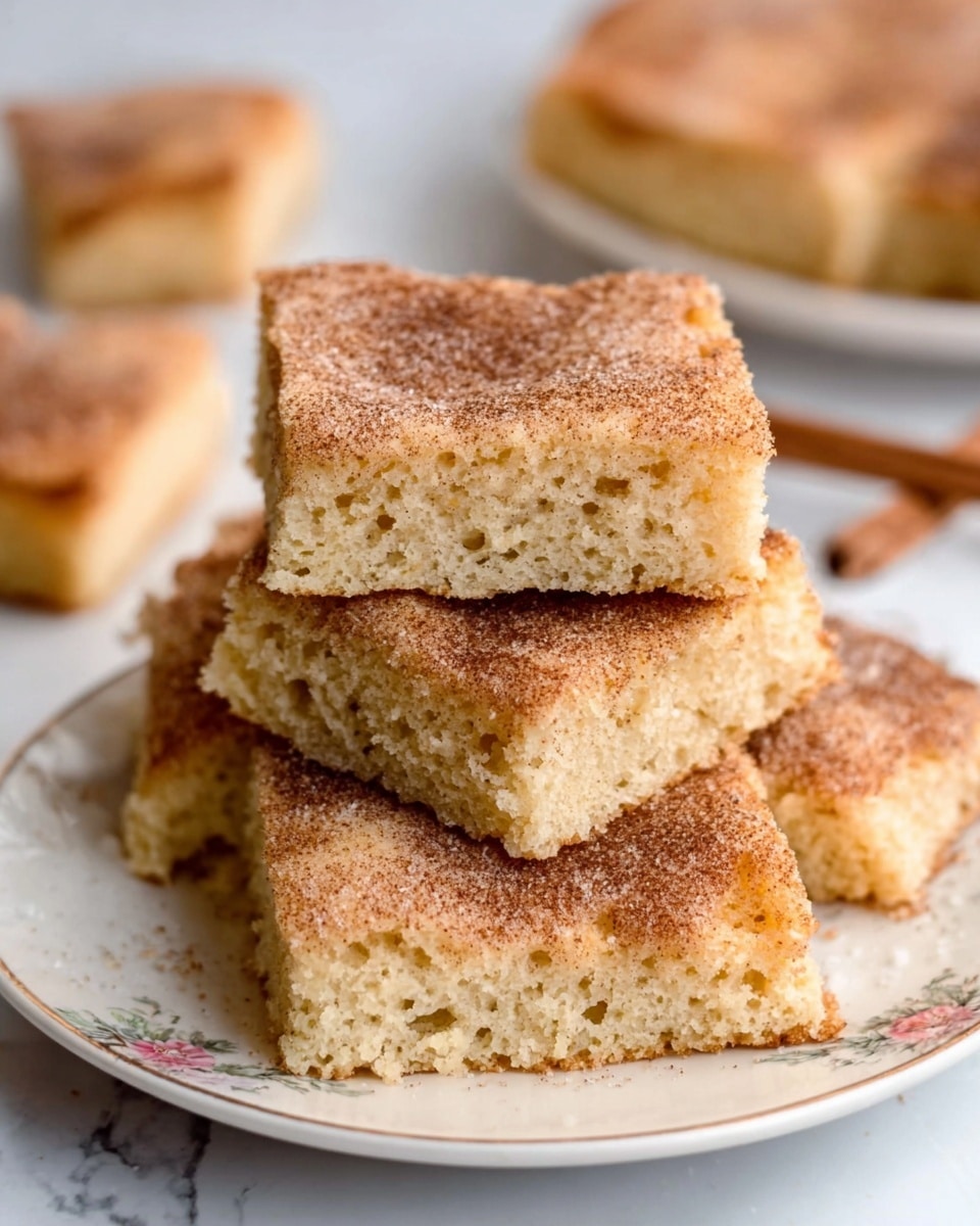 The image shows three squares of a moist cake stacked on a white plate with a subtle floral pattern. Each square has a soft, crumbly texture with a golden-brown color. The top surface of each square is coated with a layer of cinnamon sugar, giving it a slightly grainy texture and a warm reddish-brown color. The cake layers appear thick and porous, suggesting a tender inside, with the top layer slightly crisp from the cinnamon sugar. The background is a white marbled texture providing a clean and bright setting. photo taken with an iphone --ar 4:5 --v 7