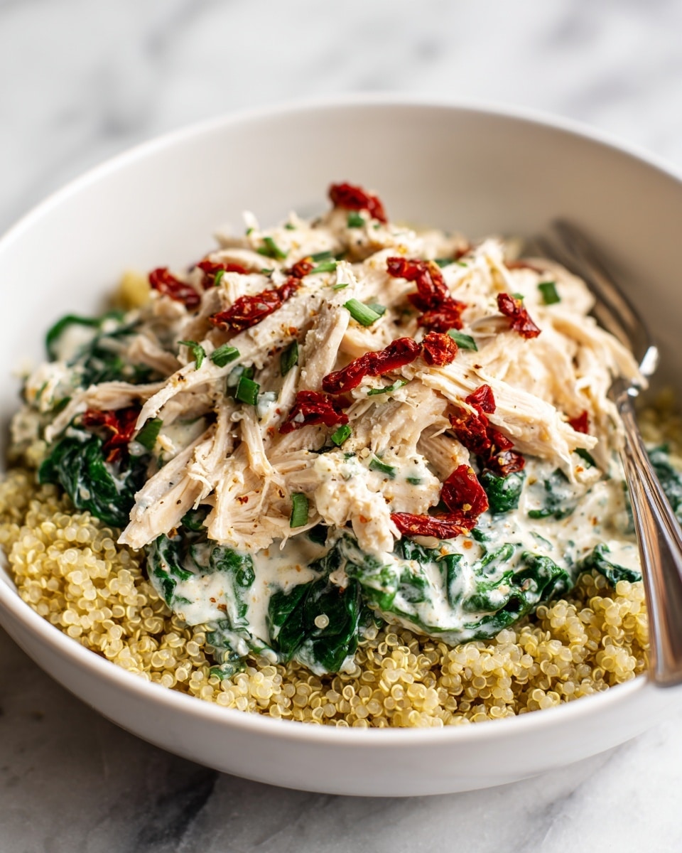 A bowl of cooked light yellow quinoa forms the bottom layer, with a creamy white sauce mixed with dark green spinach leaves resting on top. On the creamy layer, there is shredded light beige chicken scattered with small pieces of bright red sun-dried tomatoes and sprinkled with chopped green herbs. A silver fork is placed on the right side inside the bowl, which is white with a thin brown rim. The bowl sits on a white marbled surface with a soft beige cloth near it. photo taken with an iphone --ar 4:5 --v 7
