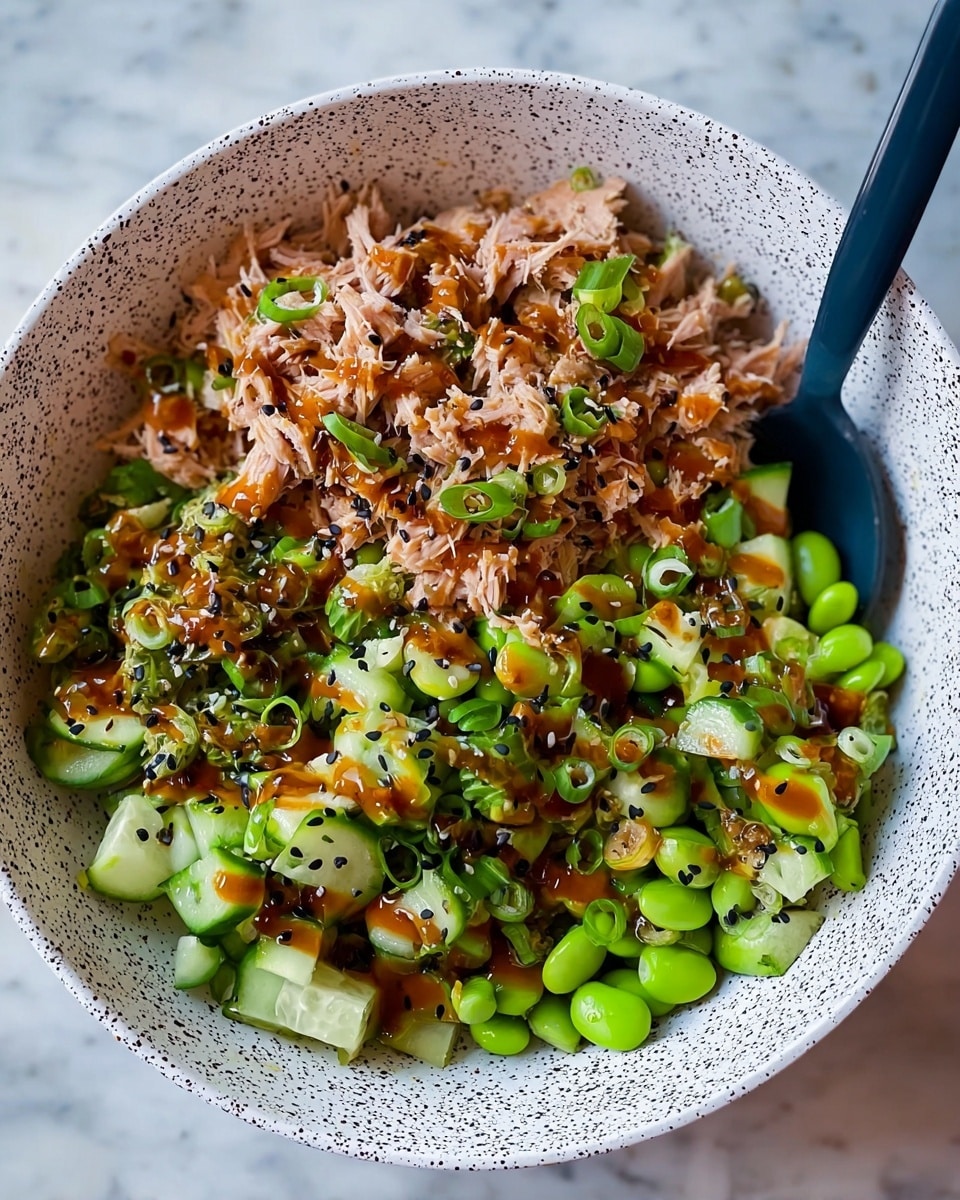 A white bowl with black speckles holds a layered dish consisting of shredded light brown tuna meat on top, surrounded by chopped bright green cucumbers and edamame beans. There is a layer of shiny brown sauce drizzled unevenly across the ingredients, sprinkled with small black sesame seeds and finely chopped green onions. The textures vary from soft tuna to crisp cucumber slices and firm edamame. A dark blue spoon rests inside the bowl on the right side. The bowl is placed on a white marbled textured surface. photo taken with an iphone --ar 4:5 --v 7