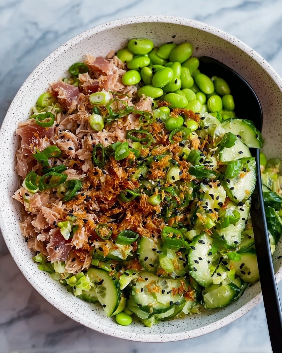 A white speckled bowl filled with a salad made of several mixed layers: shredded light pink tuna chunks, bright green edamame beans, sliced fresh cucumber pieces with dark green skin, chopped light green spring onions, and small bits of toasted brown fried onions sprinkled on top with black sesame seeds scattered over everything. A black spoon is partially inserted into the salad on the right side of the bowl. The bowl sits on a white marbled texture. photo taken with an iphone --ar 4:5 --v 7